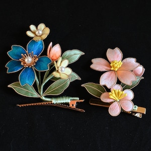 May include: Two floral hair clips. One clip features blue flowers with pearl centers and green leaves. The other clip has pink flowers with yellow centers and green leaves. Both clips have gold-colored metal clasps.