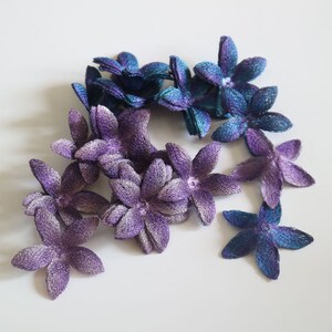 May include: A collection of purple and blue fabric flowers with a shimmery finish. The flowers are arranged in a cluster and have a delicate, feminine appearance.