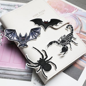 May include: Four black embroidered iron-on patches featuring a spider, a scorpion, and two bats. The patches are on a white notebook with the text "Time Flies" and "Journey" visible.