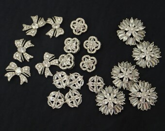 Pode incluir: Conjunto de enfeites de strass prateados em várias formas, incluindo laços, flores e designs geométricos. Os enfeites estão dispostos sobre um fundo preto.