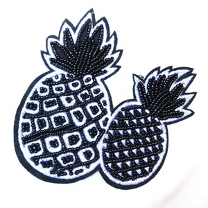 Può includere: Due toppe di ananas nere con perline e contorni bianchi. Gli ananas sono in un gruppo, uno sopra l'altro.