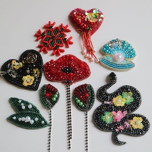 May include: A collection of beaded and sequined appliques in various shapes and colors, including a heart, a snowflake, a flower, a seashell, a snake, and a pair of leaves. The appliques are all handmade and feature intricate details.