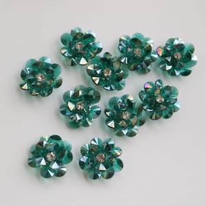 以下が含まれることがあります： 10個のティール色の花型装飾品。イリデッセントなファセットと透明なラインストーンの中心部があります。花の模様は、複数の丸みを帯びた花びらでできています。装飾品は白い表面に散らばっています。