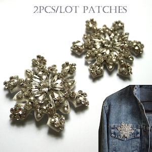 Könnte beinhalten: Zwei silberne Schneeflocken-Patches mit klaren Strasssteinen. Die Patches sind auf weißem Hintergrund und auf einer Jeansjacke abgebildet.