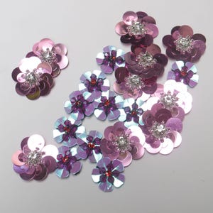 Może przedstawiać: Kolekcja aplikacji kwiatowych z cekinami w odcieniach różu i fioletu. Każdy kwiat ma centralny kryształ górski lub czerwoną szlachetną kamień. Cekiny tworzą efekt połysku, idealny do ozdabiania odzieży lub rękodzieła.