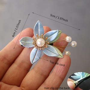 Puede incluir: Pinza para el pelo con forma de flor azul claro y verde con un centro de perla y detalles de perlas. La pinza para el pelo mide 5 cm de largo y 4 cm de ancho. La parte trasera es una pinza para el pelo.