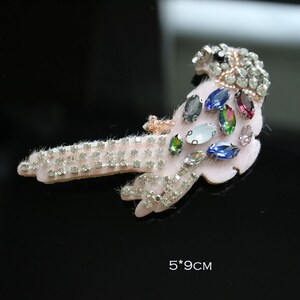 Puede incluir: Broche en forma de pájaro rosa claro, adornado con pedrería transparente y piedras preciosas de colores. La broche mide 5 x 9 cm. El cuerpo del pájaro está cubierto de pequeños brillantes brillantes y las alas presentan una variedad de gemas de colores.