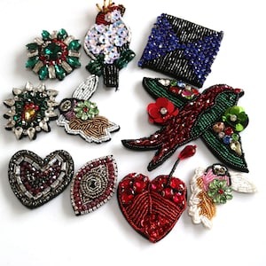 Könnte beinhalten: Sortiment an bunten, perlenbesetzten Bügelbildern. Die Patches zeigen verschiedene Designs, darunter einen Vogel, ein Herz, ein Auge, eine Blume und eine geometrische Form. Die Patches sind mit Pailletten, Perlen und Strasssteinen verziert.