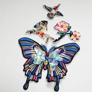 Könnte beinhalten: Gestickte Aufnäher mit einem Schmetterling, zwei Vögeln und einer Blume. Der Schmetterling hat blaue, rosa und weiße Flügel mit schillernden Pailletten. Die Vögel und die Blume haben ebenfalls farbenfrohe Pailletten-Details. Ideal zum Aufnähen auf Kleidung.
