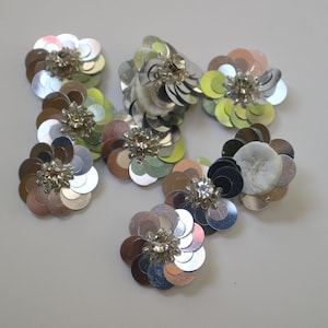 Puede incluir: Una colección de ocho adornos de flores de lentejuelas plateadas. Cada flor está hecha de lentejuelas superpuestas en un patrón circular con un centro de pedrería.