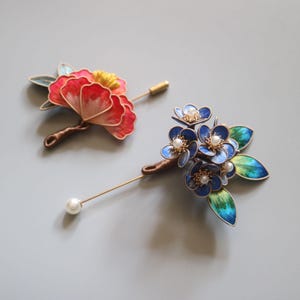 May include: Two floral brooches with pearl accents. One brooch features a red and yellow flower with green leaves. The other brooch features a cluster of blue flowers with green leaves.