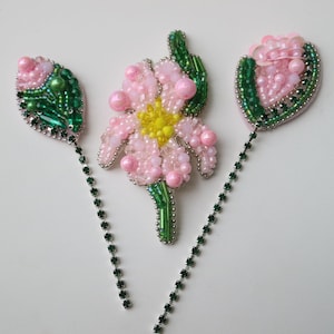 May include: Three beaded flower brooches with green leaves and stems. The brooches are pink and white with a yellow center. The brooches are attached to green chains.