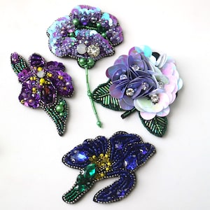 Puede incluir: Cuatro aplicaciones de flores bordadas en tonos de morado, azul y verde. Las flores están hechas con lentejuelas, cuentas y pedrería.