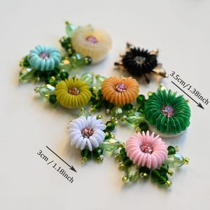 May include: A selection of decorative flower embellishments in various colours. Each flower measures approximately 3 cm to 3.5 cm in diameter. The flowers feature colours such as white, pink, green, yellow, and orange, with a central gem and green beaded accents.