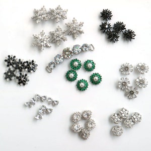 Op de afbeelding: Assortiment van zilveren en zwarte strassversieringen, waaronder bloemen, sterren, strikken en een ketting met heldere strasssteentjes. Er zijn ook groene strassversieringen met een parel in het midden.