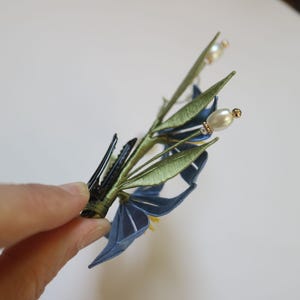 May include: A blue and green silk flower hair clip with pearl accents. The clip is attached to a green stem with leaves and has a blue flower with a yellow center.