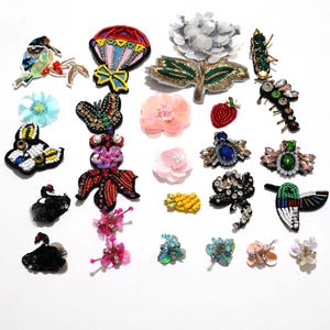 May include: A collection of colorful, beaded and sequined appliques. Designs include a bird, a hot air balloon, flowers, a butterfly, a strawberry, a pineapple, and other decorative elements. The appliques are in various colors, including red, blue, green, and pink.