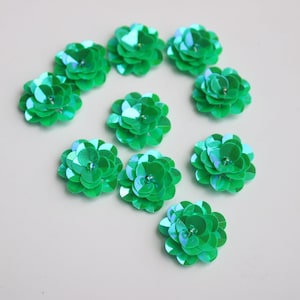 May include: Ten green iridescent flower-shaped sequins. Each sequin is made of multiple layers of overlapping sequins, creating a 3D effect.