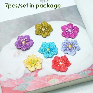 May include: Seven colourful embroidered flower patches in various colours including purple, blue, yellow, green, pink, and red. The patches are arranged on a white surface with a floral pattern. 7pcs/set in package.
