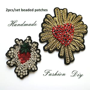 May include: Two handmade beaded patches, one with a red heart surrounded by gold beads and the other with a red strawberry surrounded by clear beads. The patches are on a white background with the text "2pcs/set beaded patches" and "Handmade" above the patches and "Fashion DIY" below the patches.