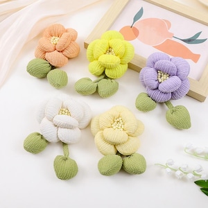 May include: Five fabric flower hair clips in various colors, including yellow, orange, white, purple, and light yellow. Each flower has a three-dimensional, layered design and is attached to a green fabric stem with two leaves.