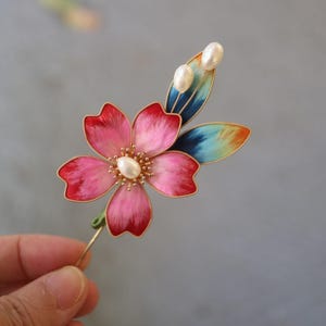 May include: A pink flower hair clip with gold wire detailing and three white pearl accents. The flower has a blue and green leaf with a rainbow gradient.