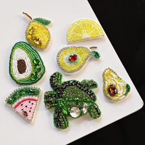 May include: A collection of beaded fruit and animal pins. The pins include a lemon, avocado, watermelon slice, and a turtle. The pins are made with colorful beads and have a detailed design. The pins are on a white surface.