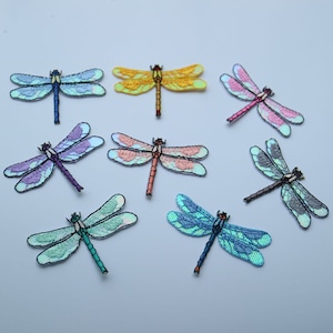 May include: Assortment of ten iridescent dragonfly iron-on patches in various colours, including blue, green, purple, pink, and yellow. The patches are perfect for adding a touch of whimsy to clothing, bags, or other fabric items.