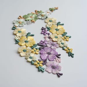 May include: Embroidered floral appliques in shades of yellow, purple, green, and white. The appliques feature detailed stitching and are arranged in a flowing, decorative pattern. Ideal for embellishing clothing or craft projects.