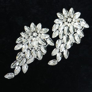 May include: Two silver floral embellishments with crystal-like accents. Each embellishment features a central flower with smaller leaves cascading down.