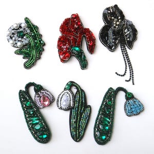 May include: Six embroidered flower patches with various colors and designs. The patches include a white flower with green leaves, a red flower with green leaves, a black flower with black leaves and a black tassel, a pink flower with green leaves, a white flower with green leaves, and a blue flower with green leaves.