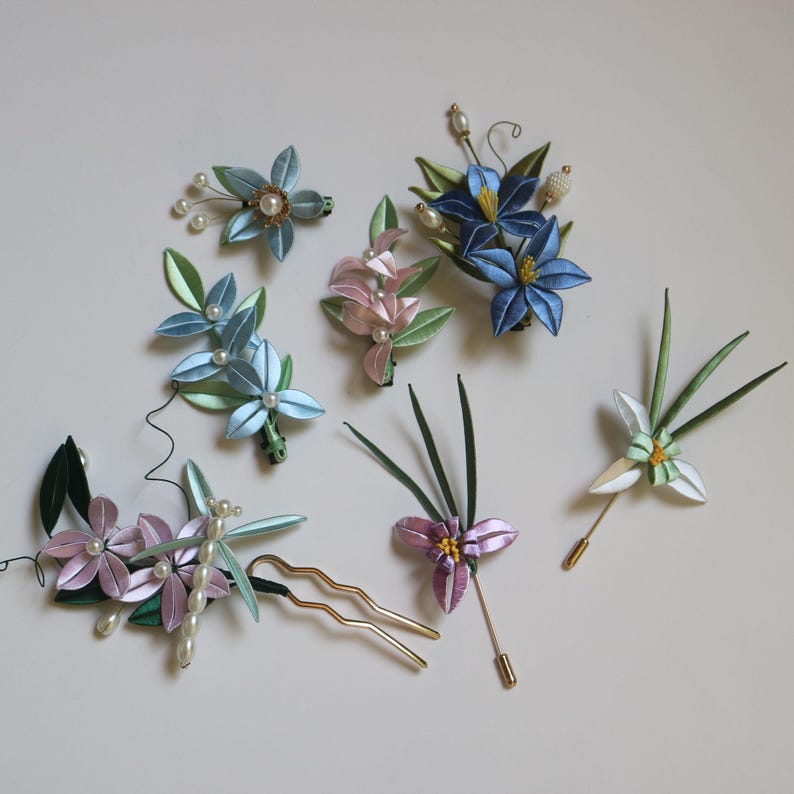 Puede incluir: Una colecci&oacute;n de accesorios para el cabello florales hechos a mano, que incluyen horquillas, pasadores y broches. Las piezas presentan flores de sat&eacute;n azul, rosa y blanco con detalles de perlas y hojas verdes. Los accesorios para el cabello son decorativos.