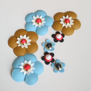 May include: An assortment of colorful flower-shaped embellishments. The flowers are in shades of blue and brown, with white and gold accents. Each flower has a red or black center, creating a vibrant and eye-catching design.