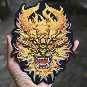 May include: Embroidered patch featuring a detailed golden dragon design with fiery accents. The dragon has an open mouth with visible teeth and is set against a black background. The patch is likely for clothing or accessories.