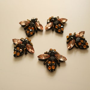 Pode incluir: Cinco enfeites em forma de abelha pretos e dourados com strass rosa e transparente. As abelhas são feitas de tecido e têm um aspeto tridimensional.