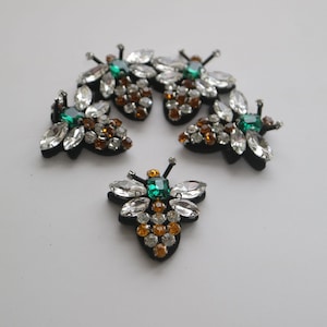 Könnte beinhalten: Fünf gestickte Bienen-Patches mit klaren und bernsteinfarbenen Strasssteinen und grünen Akzenten. Die Bienen sind auf einem schwarzen Filzgrund.