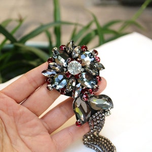 May include: A black and silver beaded embellishment with a dangling chain. The embellishment features a large, round, crystal-like center piece surrounded by smaller crystals and red beads.