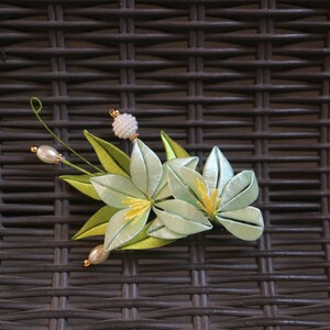 May include: A light green silk flower hair clip with yellow centers and white pearl accents. The clip is on a brown wicker background.