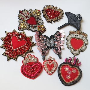 May include: Assortment of ten embroidered iron-on patches featuring hearts, butterflies, and a stingray. The patches are decorated with sequins, beads, and rhinestones in shades of red, pink, black, and gold.