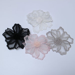 May include: Four delicate lace flower appliques in white, black, pink, and gray. Each flower has a layered design and a small center pom-pom.