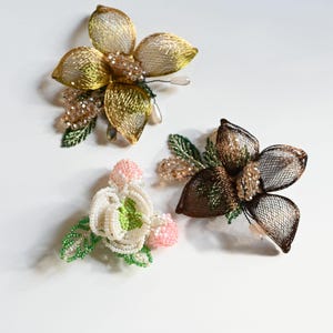 May include: Three decorative floral brooches. One is gold-toned with a mesh design and crystal accents. Another is brown with a mesh design and crystal accents. The third is white, pink, and green, made with beads.