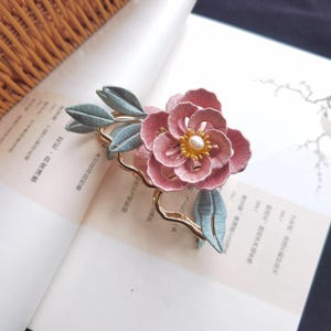 Puede incluir: Broche de flores rosas con centro de perlas y hojas azul claro. El broche está sobre una base de metal dorado. El broche está sobre un libro abierto.