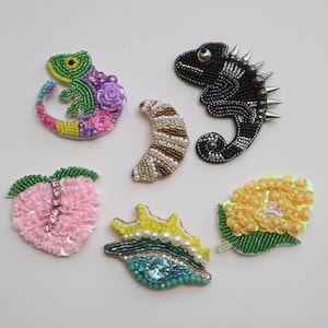 May include: A collection of six beaded patches in various shapes. Includes a green and purple chameleon, a black chameleon with silver spikes, a crescent moon, a pink strawberry, a yellow seashell, and a yellow flower.