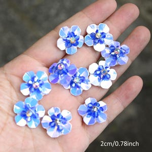 May include: A collection of iridescent blue and white flower-shaped embellishments, each with a central crystal. The flowers are approximately 2 cm in diameter. These decorative elements are ideal for craft projects.