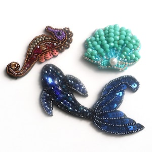 Könnte beinhalten: Drei gestickte Bügelbilder mit einem blauen Fisch, einer türkisfarbenen Muschel und einem lila-braunen Seepferdchen.
