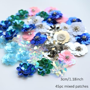 Op de afbeelding: Een verzameling van 45 gemengde patches in de vorm van bloemen. De patches zijn in verschillende kleuren, waaronder blauw, roze, groen en zilver. Elke bloem meet 3 cm. De patches zijn gemaakt van pailletten en zijn geschikt voor knutselwerk.