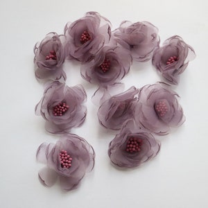 May include: A group of 12 mauve fabric flowers with pink centres. The flowers are layered and have a delicate, ruffled appearance.