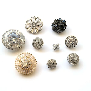 Könnte beinhalten: Eine Auswahl an dekorativen Knöpfen in verschiedenen Formen und Größen. Die Knöpfe sind mit klaren und schwarzen Strasssteinen besetzt, die in Gold- und Silbermetall gefasst sind. Zu den Designs gehören Blumen-, runde und geometrische Muster.