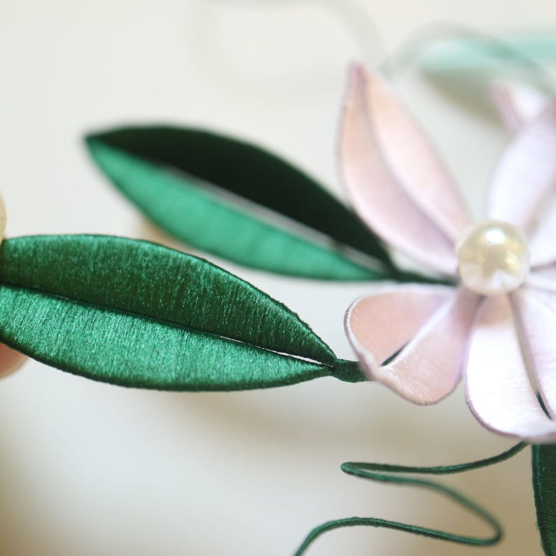 Puede incluir: Primer plano de un accesorio floral con una flor rosa claro con un centro de perla y hojas verdes. Las hojas y los p&eacute;talos de la flor tienen una textura satinada. El accesorio es probablemente hecho a mano.