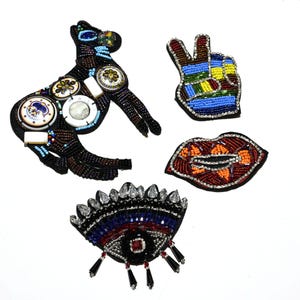 May include: Four beaded patches on a white background. The patches include a black dog, a peace sign, a pair of red lips, and an eye design. The patches are embellished with beads in various colors and shapes.